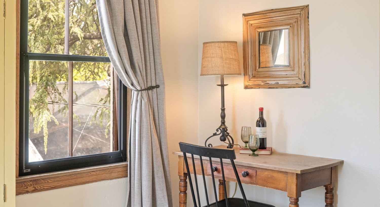 Corner of a bedroom with writing desk, black chair, lamp, bottle of wine and two glasses, near large window with grey curtain