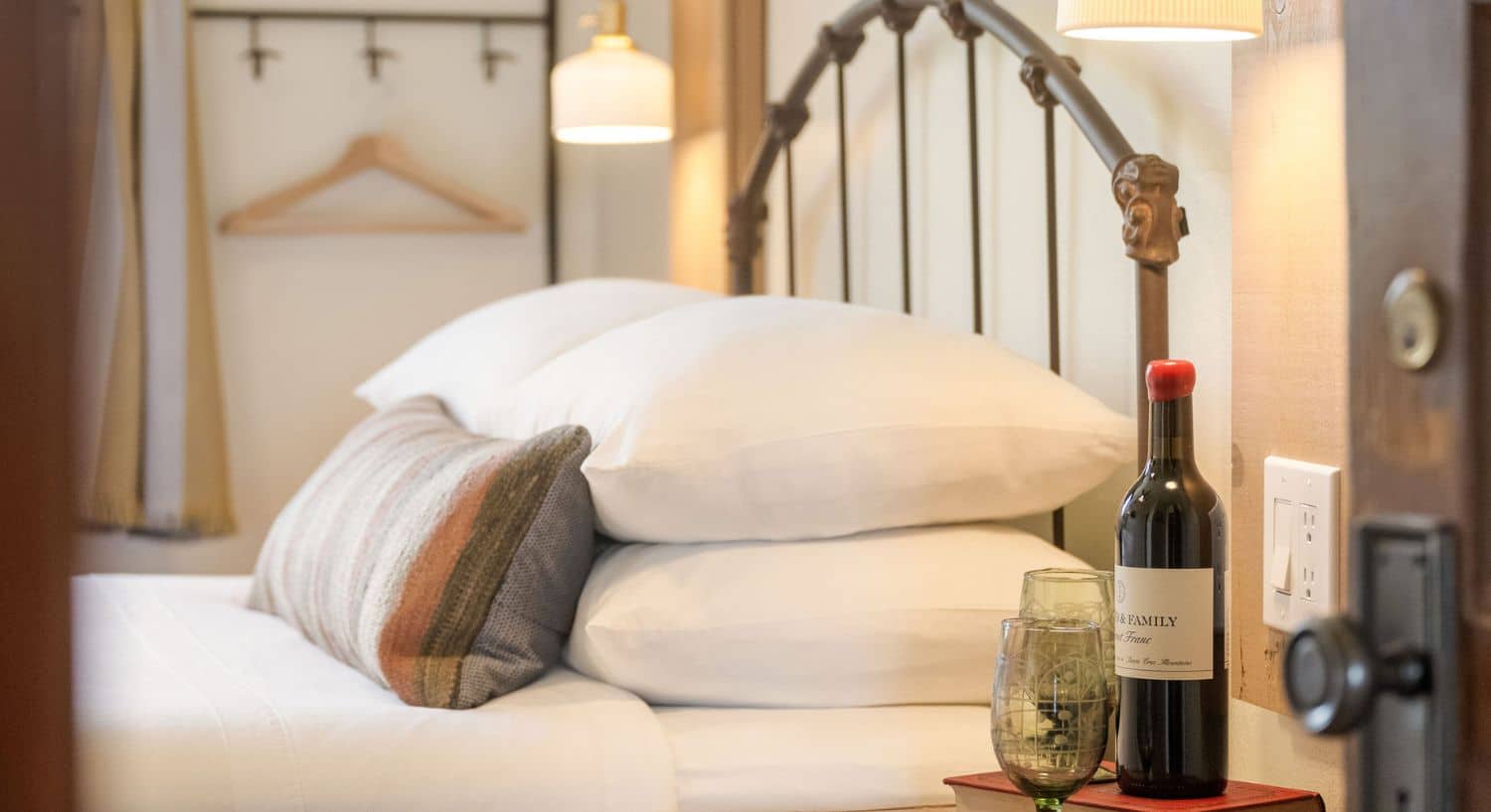 Bed with curved wrought iron headboard, plush white pillows, white linens, side table with bottle of wine and two glasses