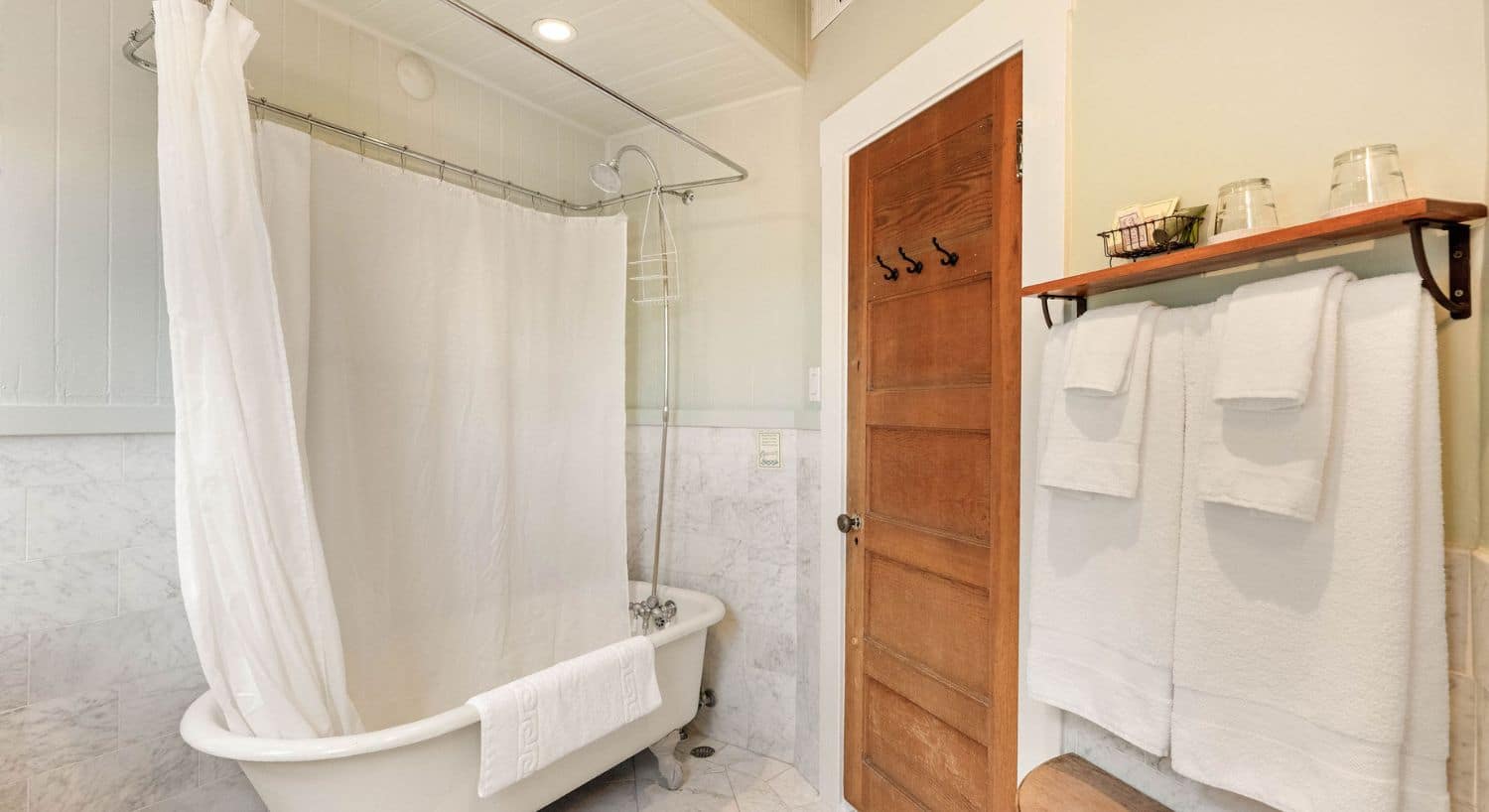 Bathroom with white clawfoot tub, white curtain, wood door and plush white towels hanging on a rack wit a wood shelf