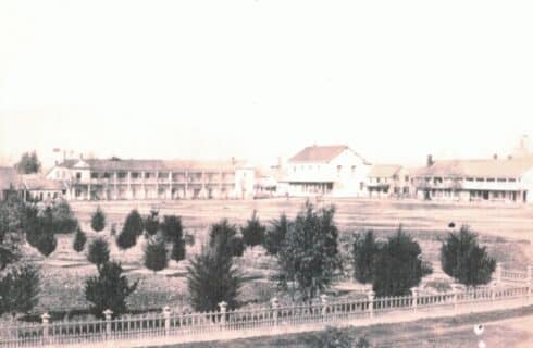 Historic black and white photograph of an expansive hotel with lots of open land and decorative fencing around a group of trees