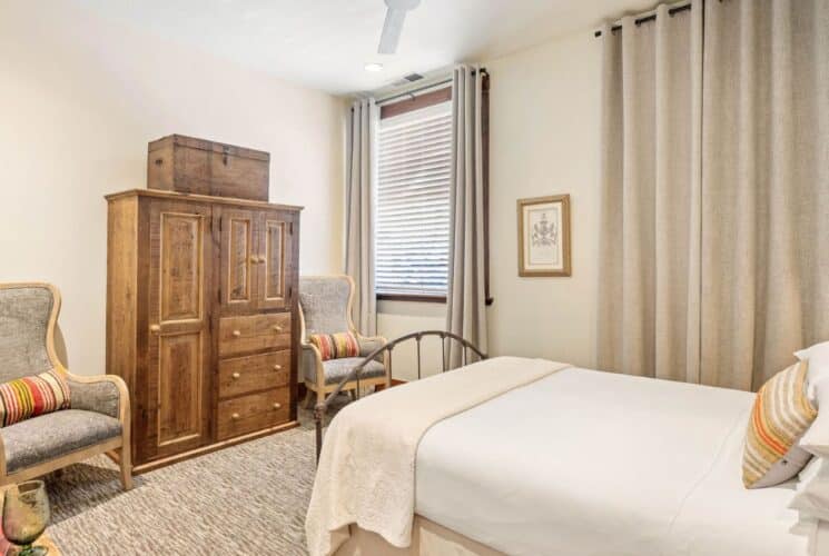 Elegant bedroom with bed in white linens, two sitting chairs, tall wood armoire and two windows with curtains