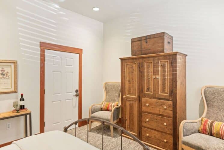 Cozy bedroom with bed, two sitting chairs, tall wood armoire and side table with bottle of wine