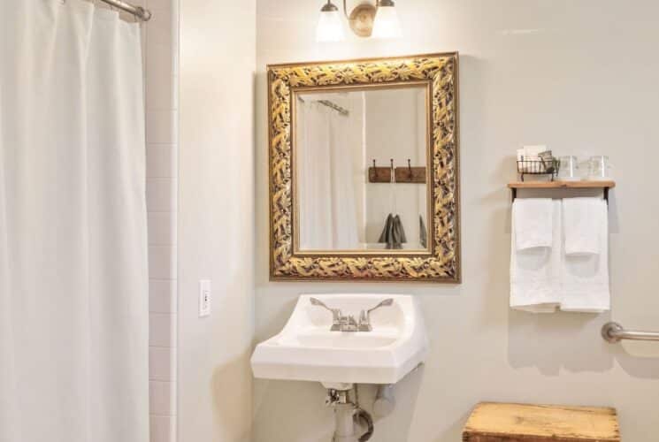 Bathroom with white shower curtain, sink, gold framed mirror and towels on a rack with a wood shelf