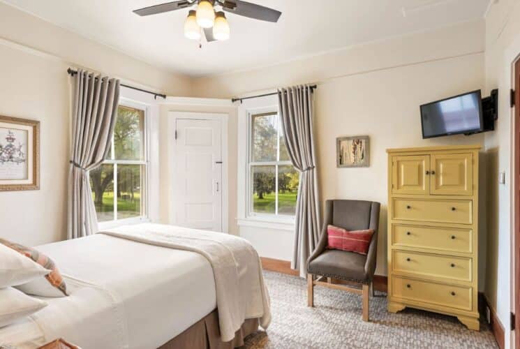 Bedroom with single bed, dresser, sitting chair, two windows with curtains