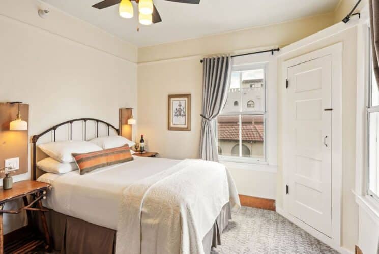 Small bedroom with queen bed, side tables with lamps, corner door and window with a curtain pulled back