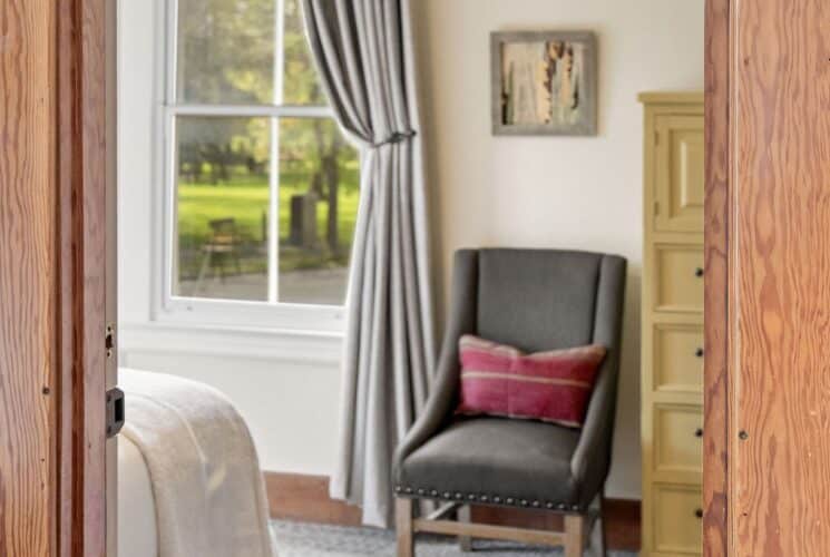 Doorway looking into a bedroom with bed, sitting chair, dresser and large window with a curtain pulled back.