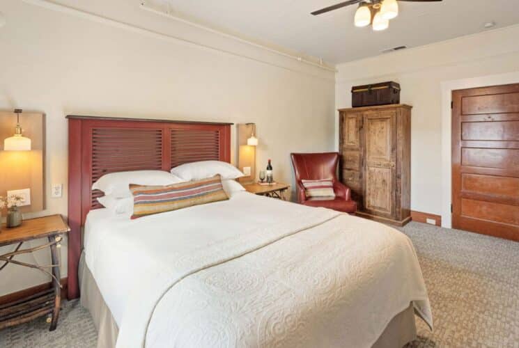 Bedroom with bed in white linens, side tables, leather sitting chair, tall wood armoire, wood doors