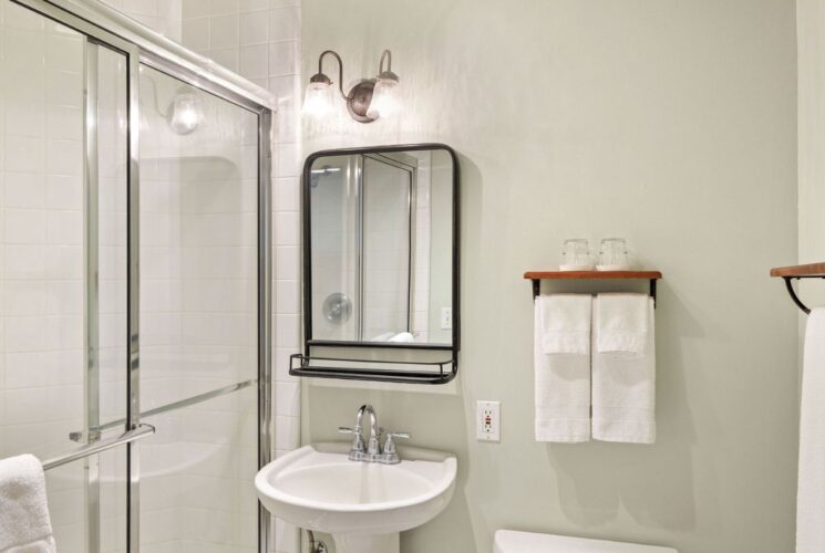 Bathroom with glass door shower, pedestal sink, black framed mirror, white towels on racks with wood shelves