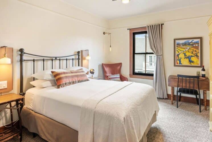 Bright bedroom with king bed, writing desk with chair, tall yellow armoire and large window with curtain