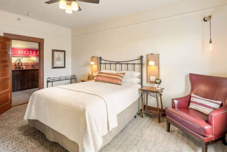 Elegant bedroom with queen bed, side tables, lamp sconces, leather sitting chair next to a bright window