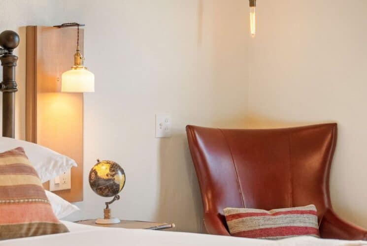 Bedroom with white bed, side table with globe, lamp sconce, leather chair with striped pillow