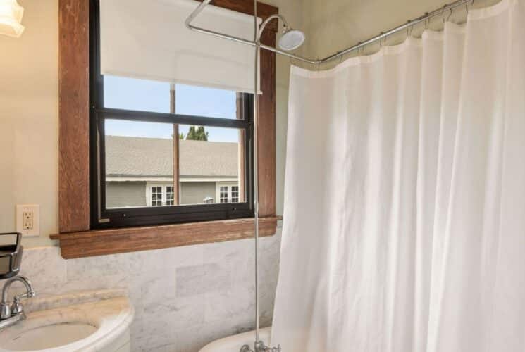 Bathroom with clawfoot tub, white shower curtain, corner sink, large window with white shade