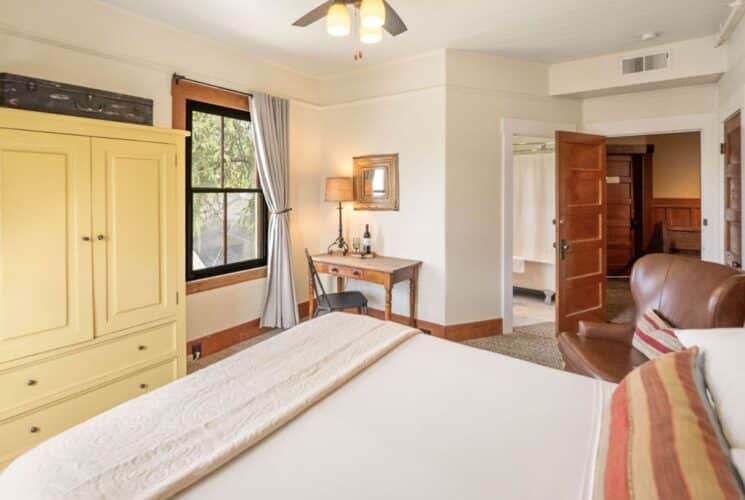 Spacious bedroom suite with bed, yellow armoire, desk and chair by a window and doorway open to a bathroom