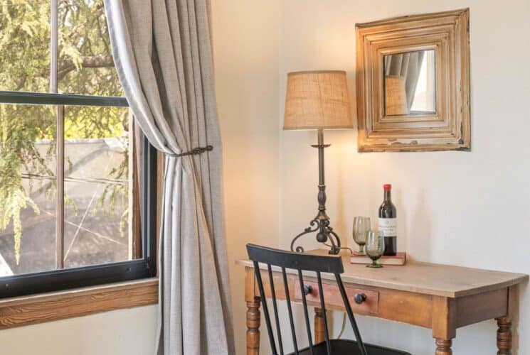 Corner of a bedroom with desk, chair, bottle of wine and two glasses and lamp next to a large window with curtain draped back