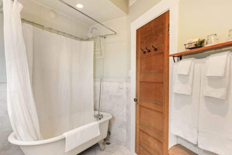 Bathroom with clawfoot tub, white shower curtain, plush towels hanging on a rack with a wood shelf