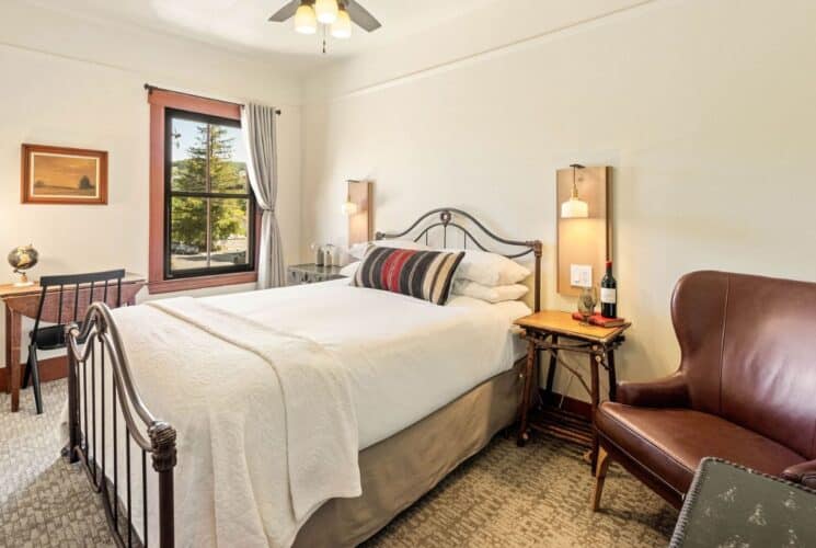 Bright bedroom with bed in white, striped throw pillow, leather sitting chair, side tables with lamps, writing desk by large window