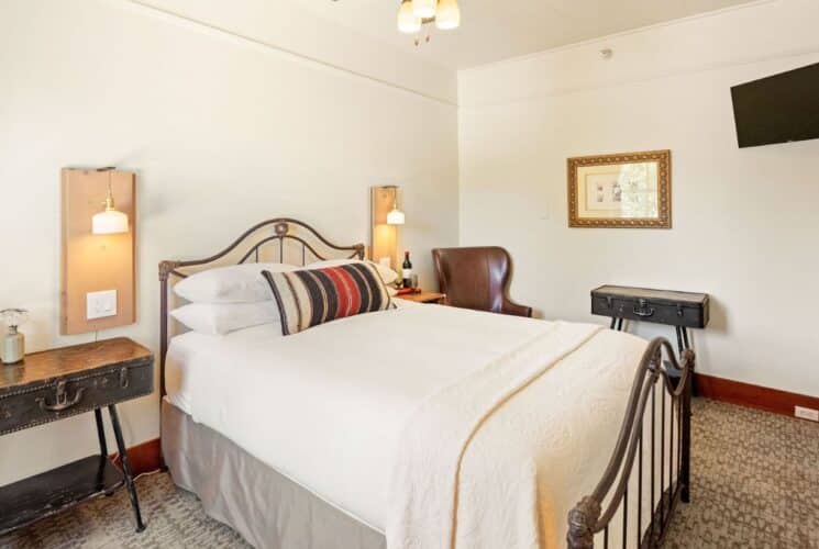 Cozy bedroom with bed in white linens, striped throw pillow, side table, leather sitting chair and corner hung TV