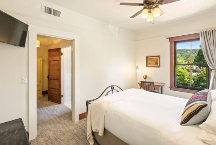 Spacious bedroom with bed in white linens, small desk with chair, TV and large window with curtains