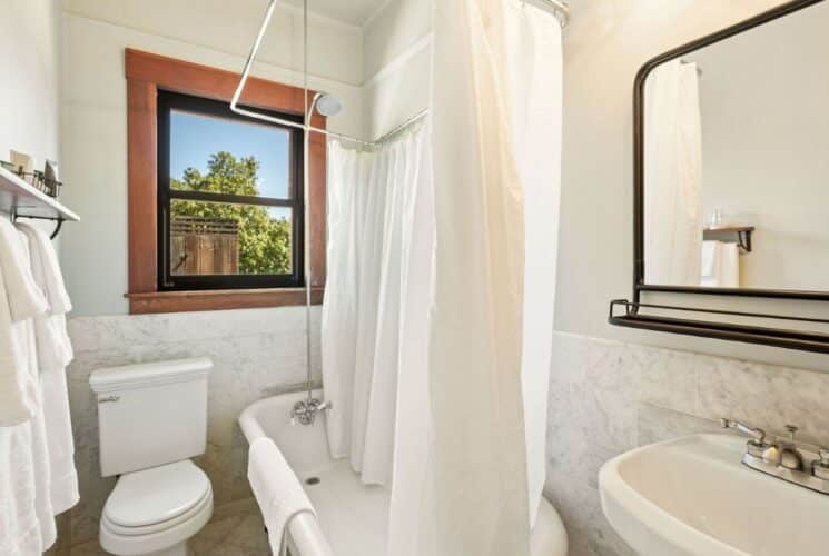 Bathroom with clawfoot tub, white shower curtain, pedestal sink, black framed mirror, plush towels and large window