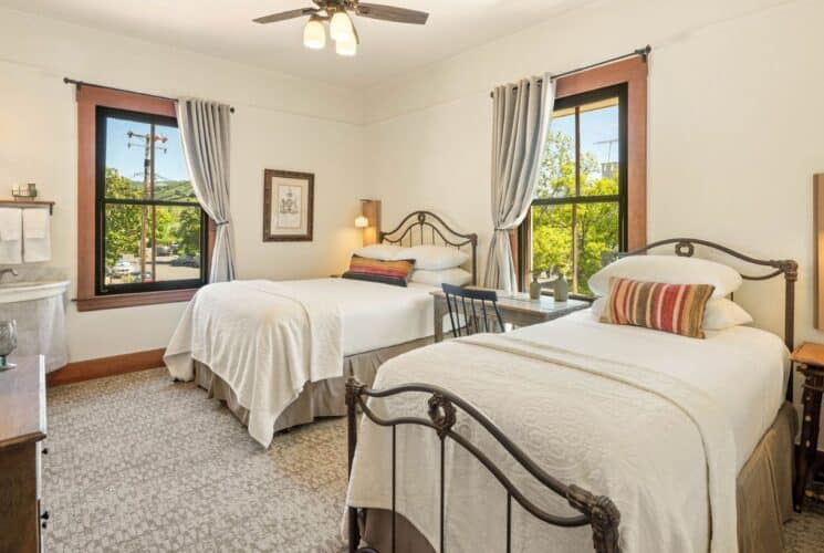 Spacious bedroom suite with two beds in white linens, writing desk and chair under a window, corner sink by a window