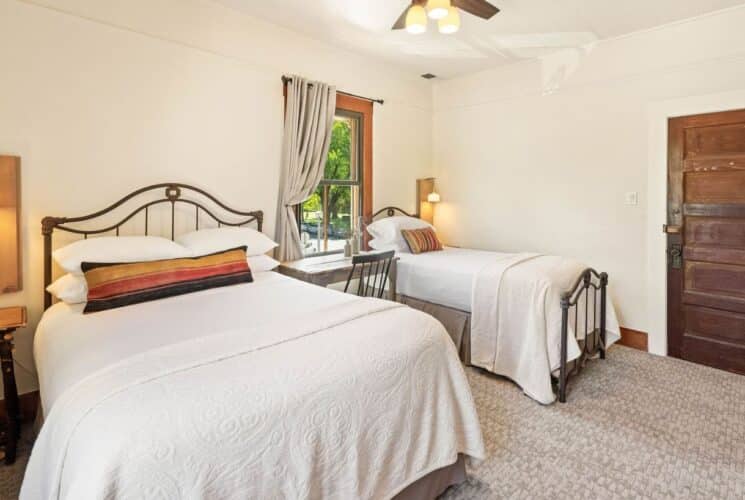 Spacious bedroom with two beds in white linens, writing desk and chair under a window, side tables with lamps