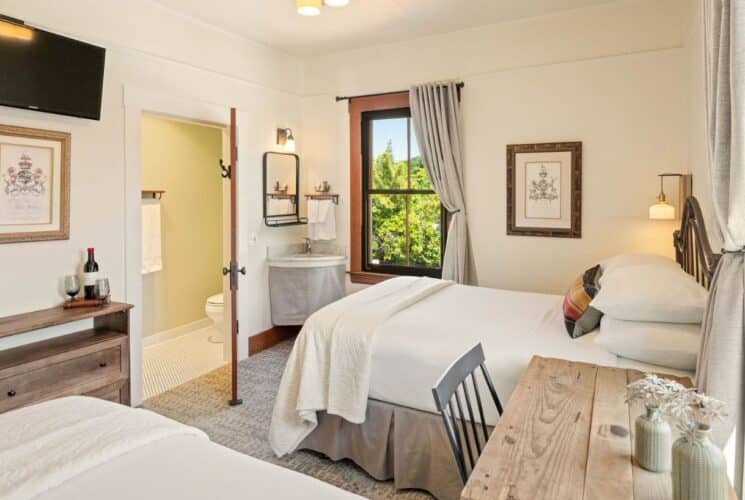 Spacious bedroom suite with two beds, writing desk and chair under a window, TV, and doorway open to a bathroom