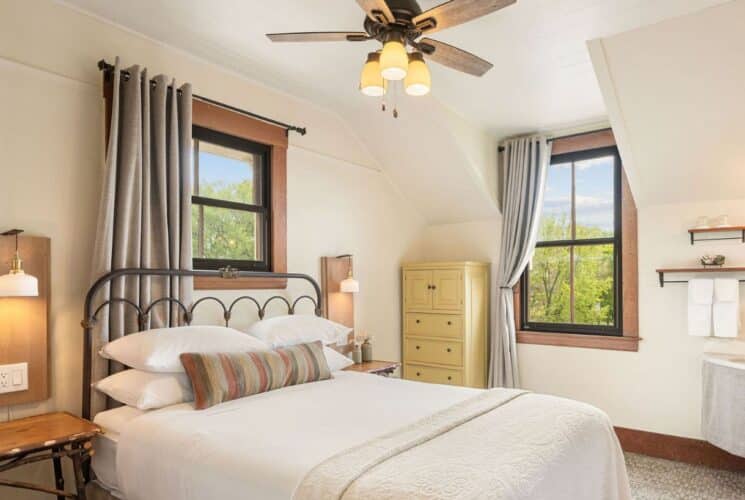 Bright bedroom with queen bed in white, corner sink, mirror, towel rack, two large windows with curtains