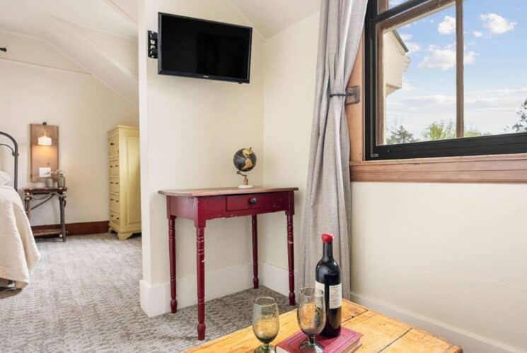 Bright bedroom suite with bed, dresser, sitting room with table with wine and glasses, TV and large windows