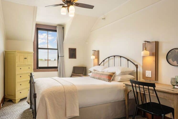 Bright bedroom with bed in white linens, tall dresser, desk with chair, bottle of wine and glasses and large window with curtains