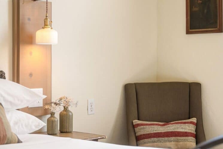 Bedroom with bed, plush pillows, side table with vases of flowers and sitting chair with a striped pillow