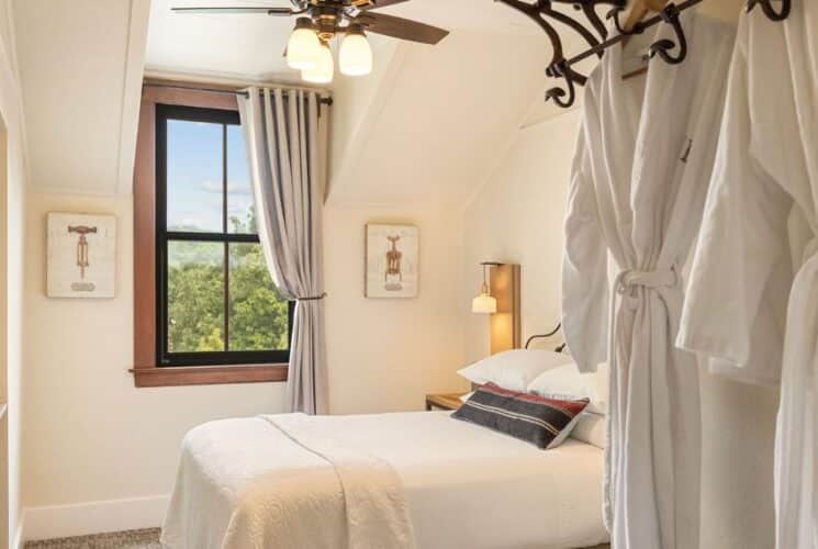 Bedroom with queen bed, window with curtain, plush white robes hanging on hooks by the door