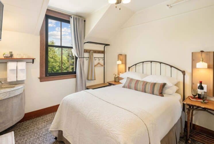 Quaint bedroom with queen bed in white, side tables with lamp sconces, corner sink with mirror, large window with grey curtain