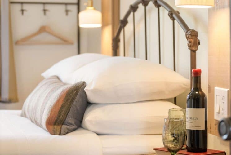 Bed with curved headboard, plush white pillows and table with bottle of wine sitting on a book with two glasses