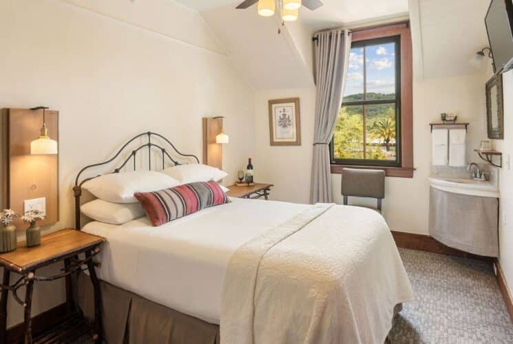 Bright bedroom with queen bed in white, side tables with lamp sconces, corner sink, large window with grey curtain