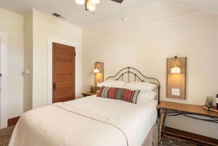 Bedroom with bed in white linens, table with bottle of wine and two gasses, lamp sconces and two wood doors