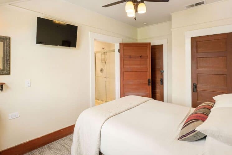 Cozy bedroom with bed in white linens, corner sink, TV and door open into a bathroom