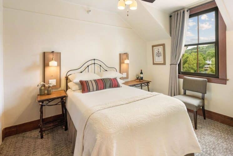 Bright bedroom with bed in white linens, side tables with lamps, sitting chair, corner sink and large window with grey curtains