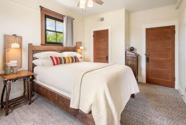 Bedroom with wood and cream tones, side tables with lamps, three wood doors and window with a curtain