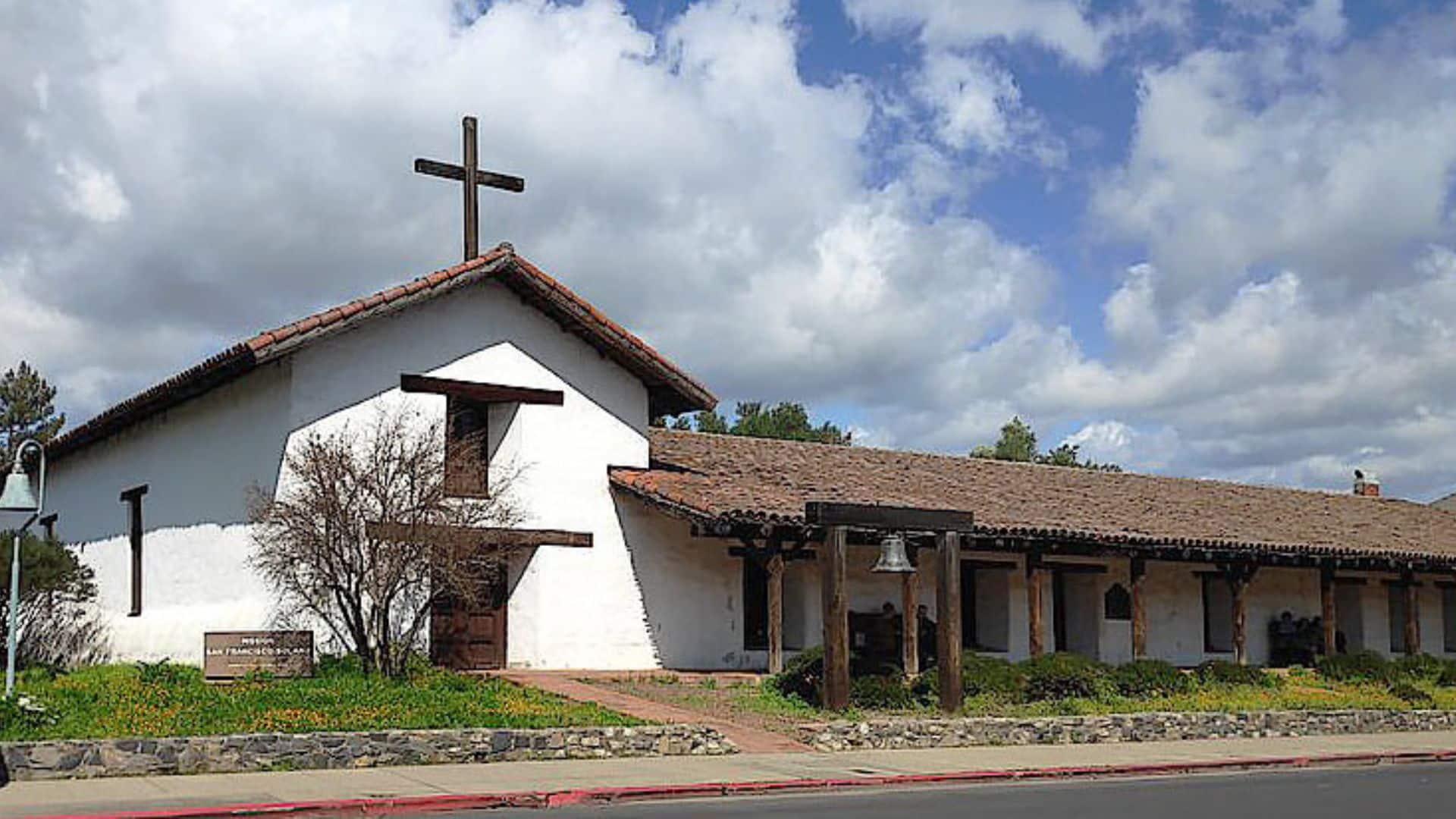 Large white building with a cross on the roof, Spanish-style shingles and arch with a bell by the walkway to the front door