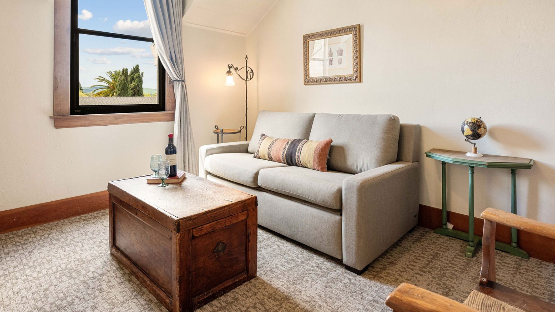 Sitting area with grey loveseat, chest, side table with globe, large window with single curtain