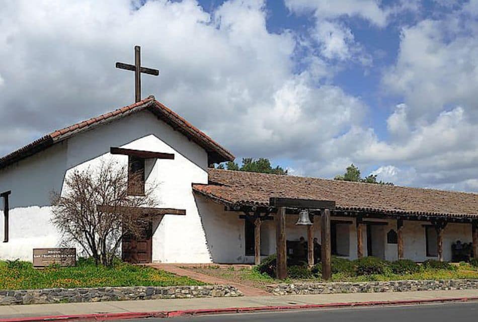 Large white building with cross on a Spanish style roof and arch with a bell by the front sidewalk