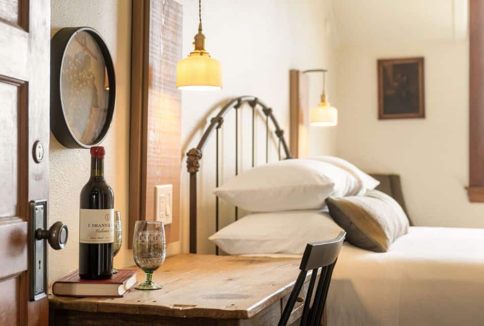 Bed with curved iron headboard, plush white pillows, desk with chair and bottle of wine on top of a book next to two glasses