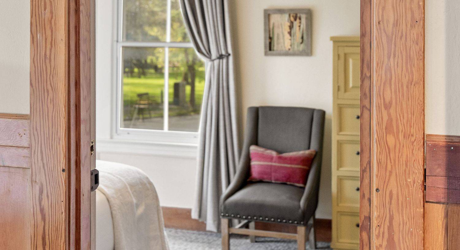 Bedroom with bed, grey sitting chair, tall dresser and large paned window with curtain